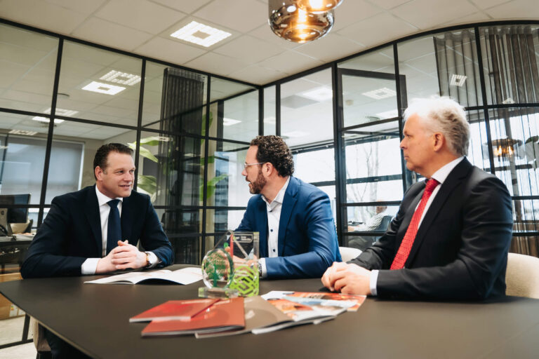 Three men in suits sit at a conference table in a glass-walled office, discussing documents.