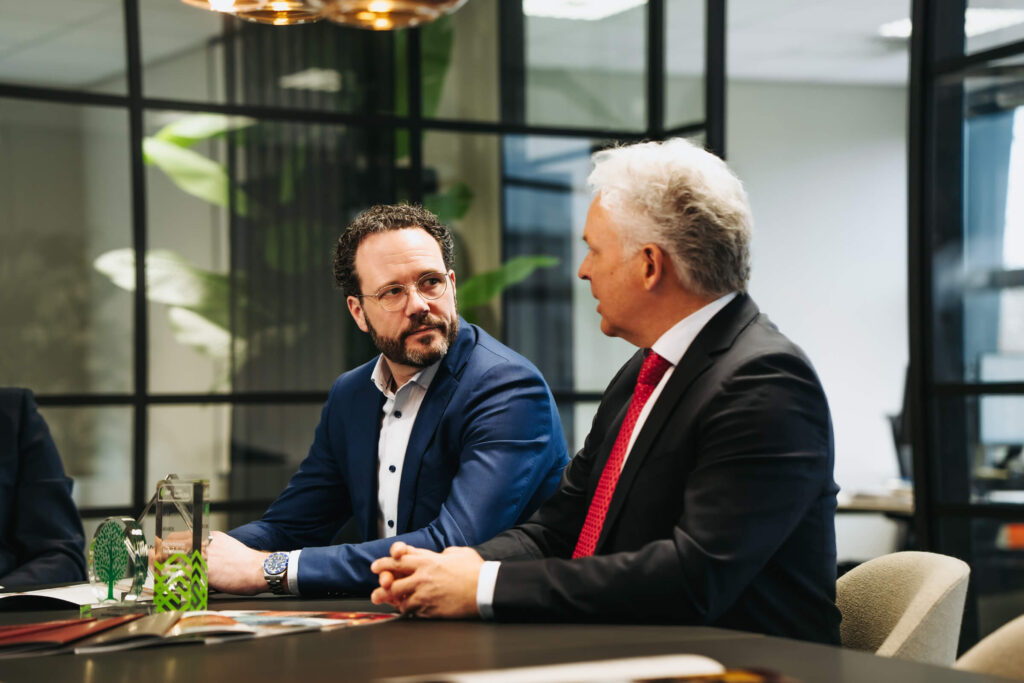 Two men in business attire sit at a conference table, engaged in conversation in a modern office with glass walls and plants in the background, trophies visible on the table in front of them.