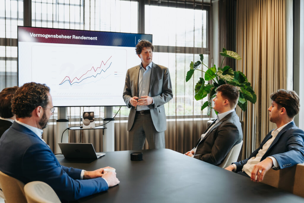 Presenter in a gray suit explains a line graph on a large screen to seated colleagues in a conference room.