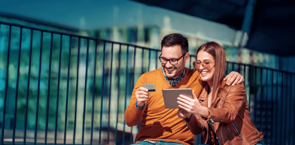 Two friends sit outdoors by a railing, smiling while sharing a tablet and a card together.