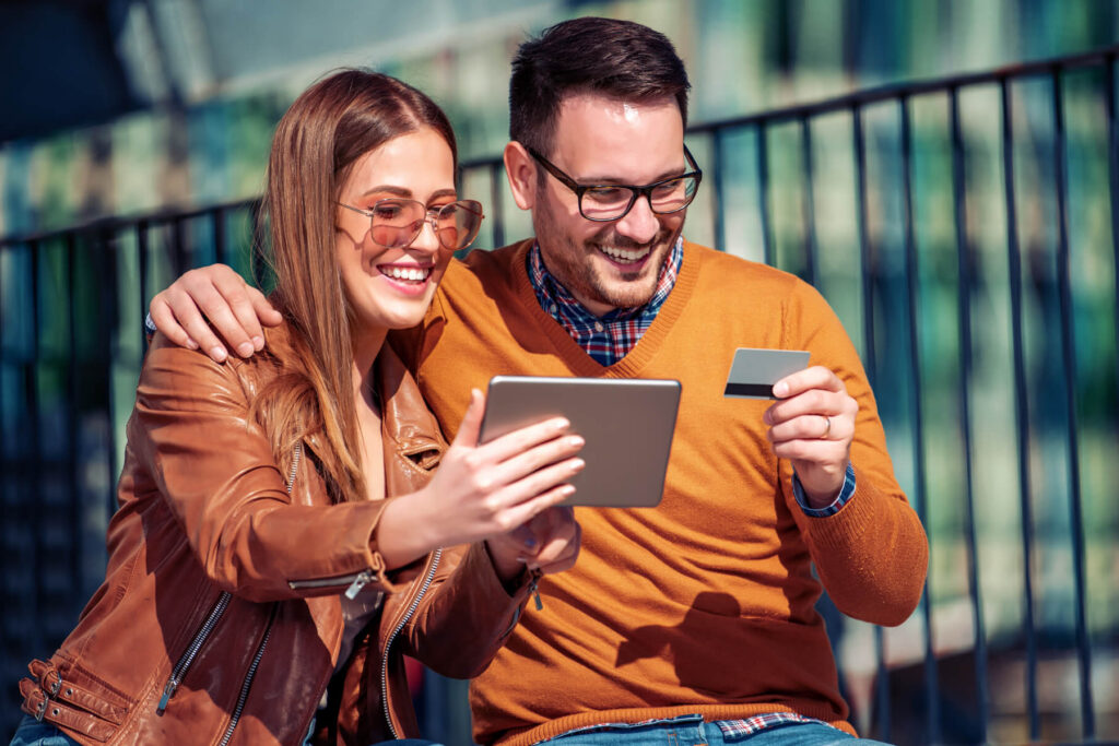 Two smiling friends sit outdoors, looking at a tablet while one holds a credit card in hand.