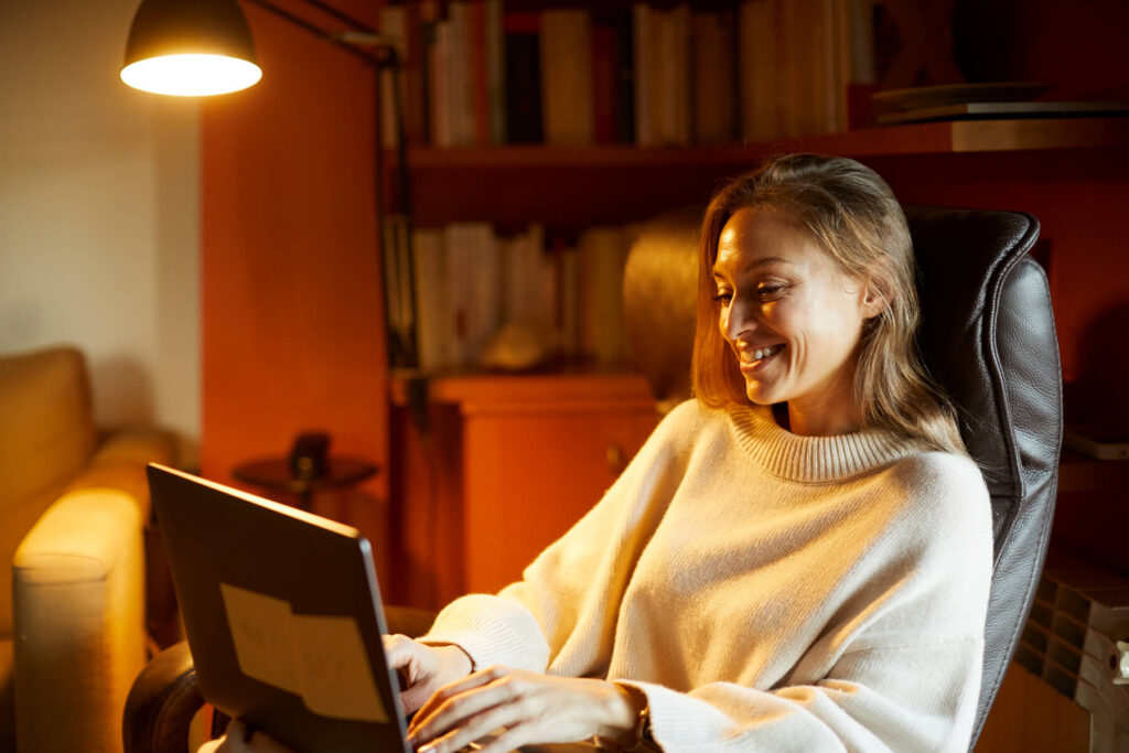 Woman smiling and typing on a laptop in a warmly lit living room.