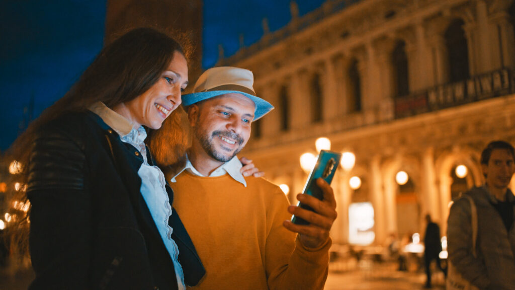 Two friends taking a nighttime selfie in a warmly lit city square, smiling at the camera.
