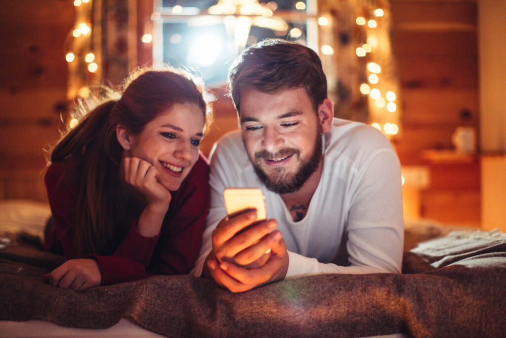 Couple lying on a bed, looking at a lit phone screen, smiling with warm string lights in the background.