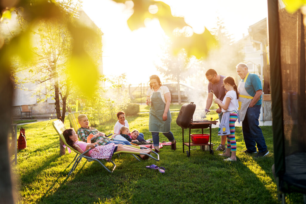 Family enjoying a sunny backyard barbecue: adults grill while kids lounge on lawn chairs nearby.