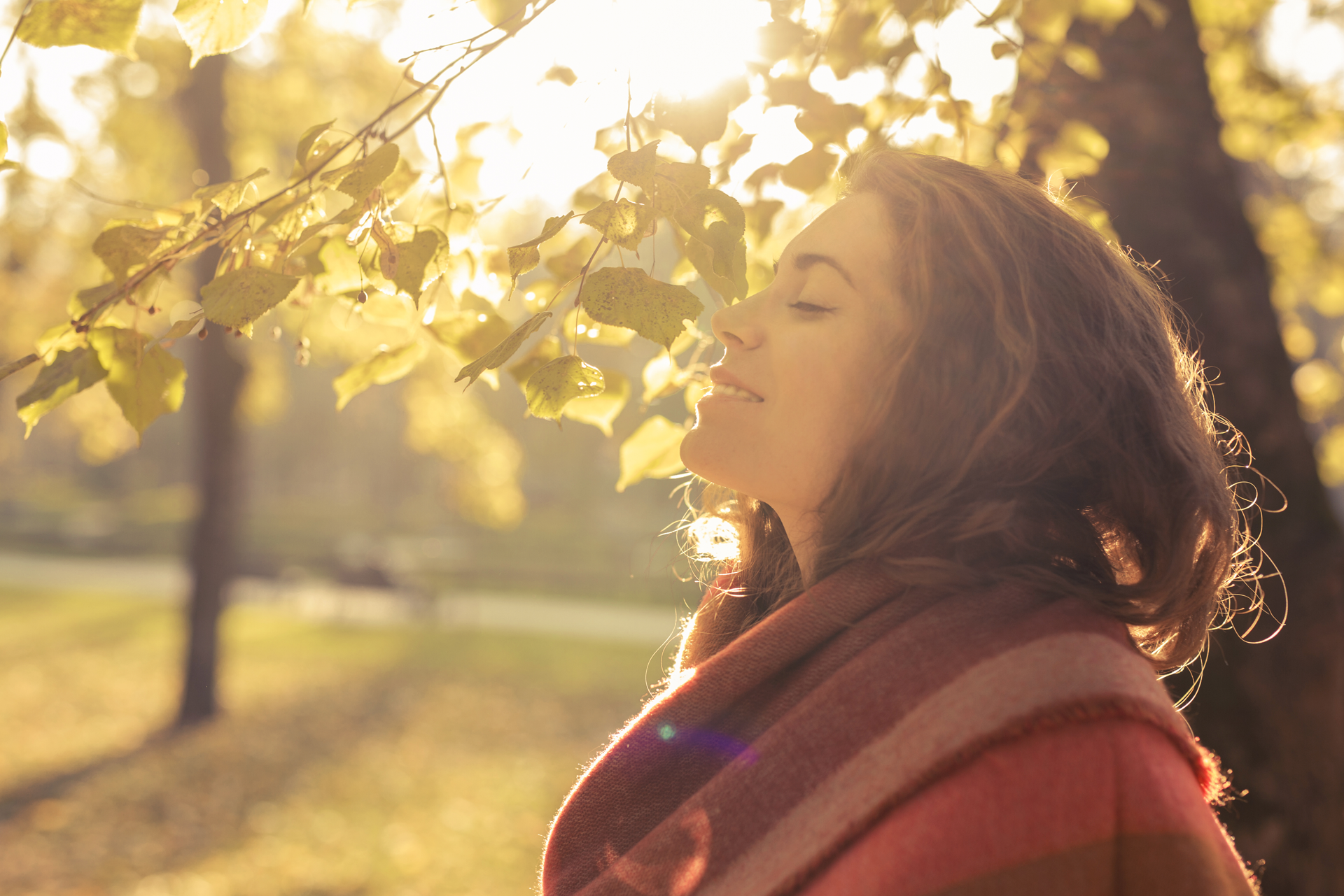 Woman in a warm scarf smiles with eyes closed as sunlit autumn leaves frame her face.