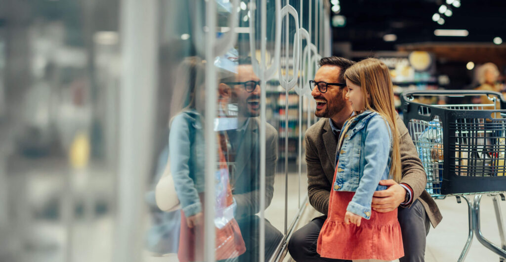 Father and daughter smile while looking at refrigerated display cases, holding onto a shopping cart nearby.