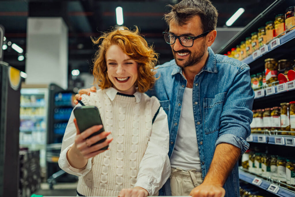 Couple in a grocery store smiling while looking at a smartphone together near refrigerated and shelf items behind them.